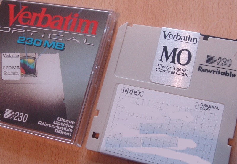 A photo of a 230MB MO Disk made by Verbatim with 230mb capacity. The drive and it's cartridge case are shown. It indicates that the disc is Rewritable and includes wording Disque Optique 90mm.
