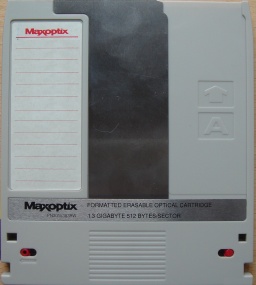 The front view of a Maxoptic Magneto Optical MO disk 1.3GB. The disk is grey colour with standard notches out of the side indicating ISO compliant formatting. The disk has it's read protect red tabs in thhe write protect position. 